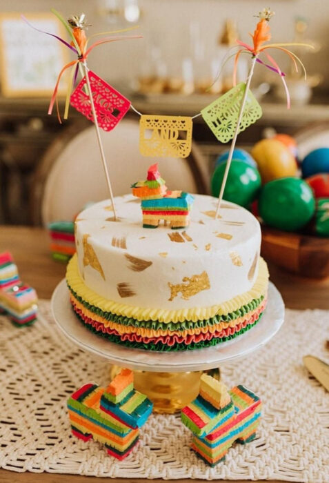 Decoración de Navidad con temática mexicana; pastel temático en la mesa, con papel picado, piñata de caballo de colores rojo, amarillo, verde, azul y rosa