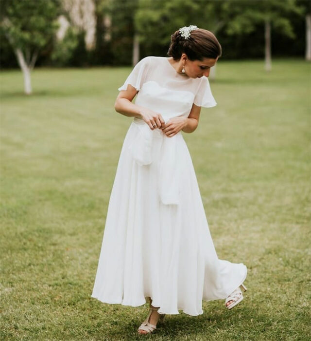 Novia en jardín con recogido elegante con peineta blanca con vestido hasta los tobillos con transparencia en la parte superior y sandalias beige de tacón 