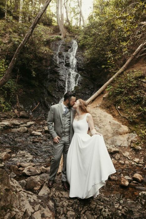 Pareja de esposos besándose en un bosque con una mini cascada entre las rocas detrás, él vistiendo un traje gris con corbata plateada y ella un vestido blanco de tirantes grueso largo hasta el piso con una media coleta