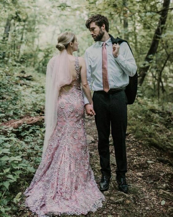 Pareja de esposos posando en el bosque, él de frente con blusa blanca, corbata rosa, pantalón negro y el saco sobre el hombro y ella con recogido bajo, velo rosa y vestido rosa corte sirena con escaje