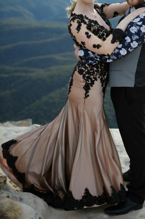 Pareja de esposos abrazándose, él con pantalón y zapatos negros y chaleco gris y ella con vestido en corte sirena color dorado con una capa transparente de encaje negro 