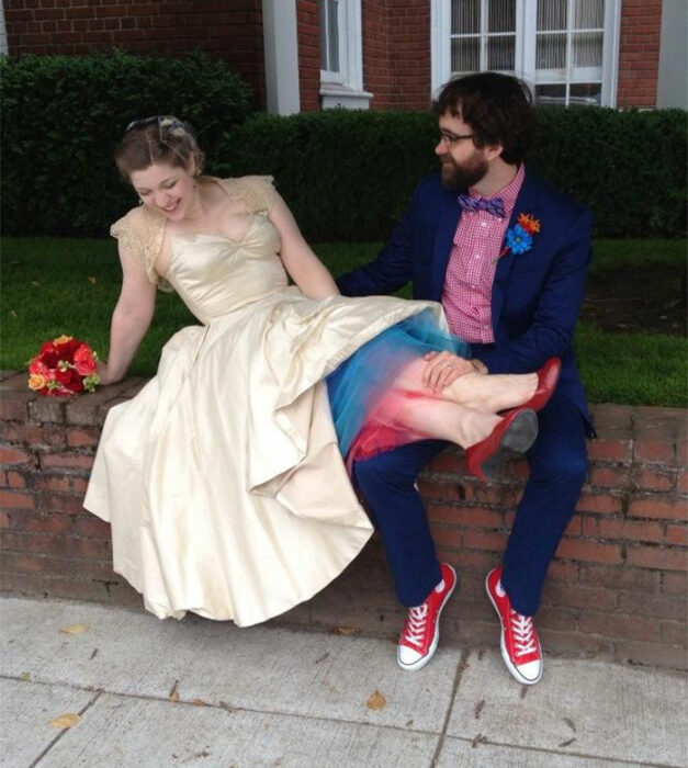 Pareja de esposos sentados en una bardita de ladrillos rojos. Él vistiendo un traje azul marino, camisa roja de cuadros blancos y converse rojos. Ella con vestido color perla con fondo azul y rojo y zapatos rojos con ramo de flores en color naranjas y rojo