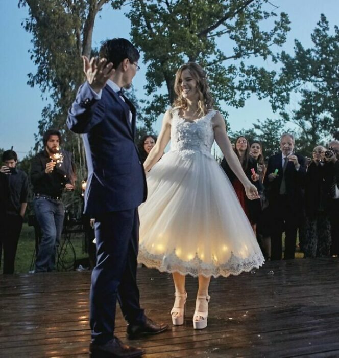 Pareja de novios baila en la pista con los invitados viéndolos al rededor; él con traje azul marino y ella con vestido de encaje hasta el tobillo con luces en la falda