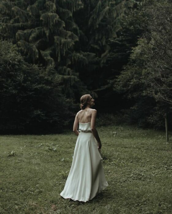 Novia de espaldas caminando por el bosque con un vestido de seda blanco hasta el piso de tirantes con un recogido bajo