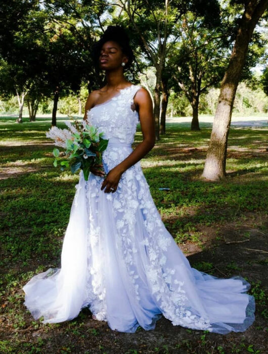 Mujer negra posando en un parque con un vestido de novia blanco, detalles de flores en 3D con solo un tirante grueso de lado izquierdo y un ramo de plantas verdes