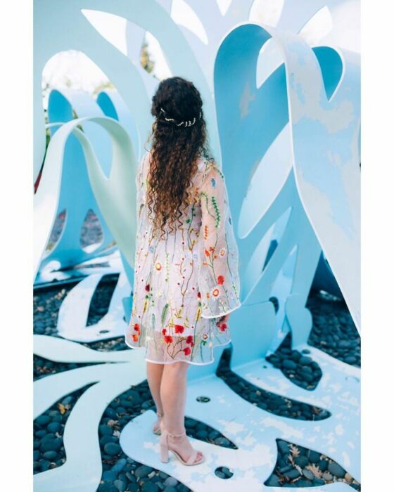 Novia con cabello rizado largo de espalda mirando hacia arriba con un vestido blanco hasta las rodillas con una capa de transparencia con flores de colores y sandalias beige de tacón