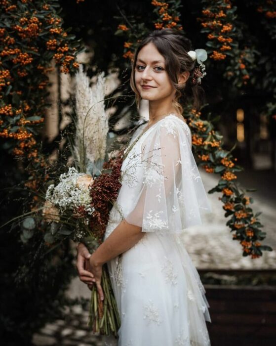 Mujer castaña con recogido de novia despeinado con vestido estilo bohemio con mangas transparente y un ramo de flores en color guinda, blanco y naranja