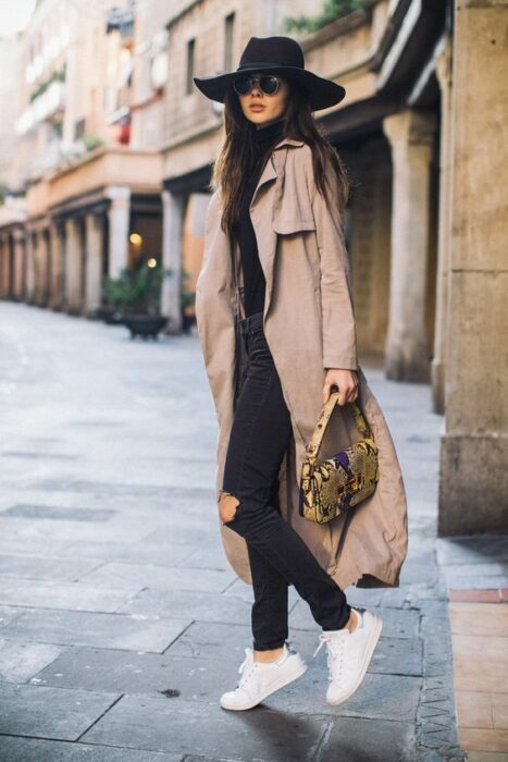 Chica usando sombrero de color negro con outfit de tenis blancos, jeans y blusa negra, junto una gabardina camel