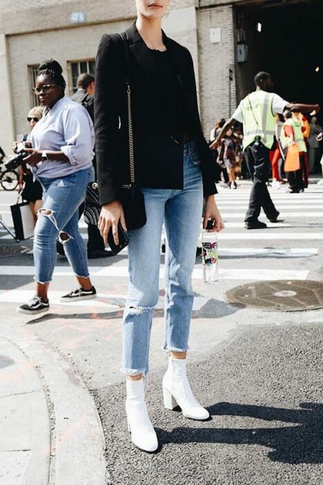 Chica usando botines blancos y jeans denim, con saco y blusa negra con bolsa de mano del mismo color