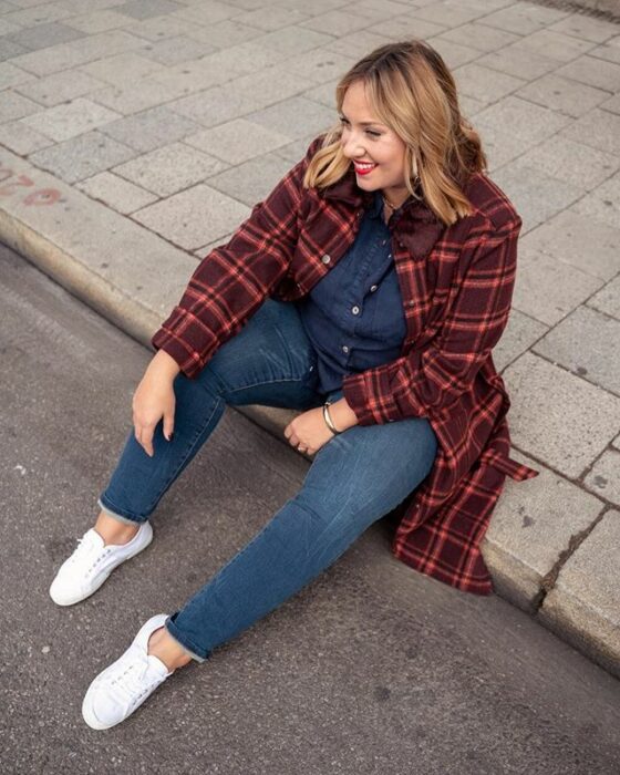 Chica curvy usando jeans y blusa denim, tenis blancos y abrigo color ladrillo a cuadros