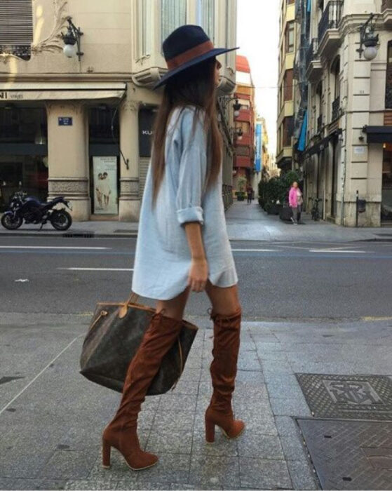 Chica usando sombrero de color negro con outfit de botas altas camel, vestido denim tipo camisa, bolso de mano color café