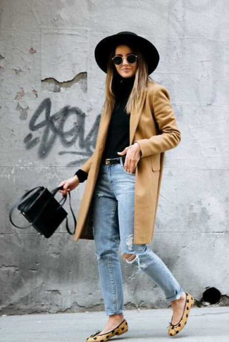 Chica usando sombrero de color negro con outfit de falts, jeans rasgados, blusa negra y gabardina color camel