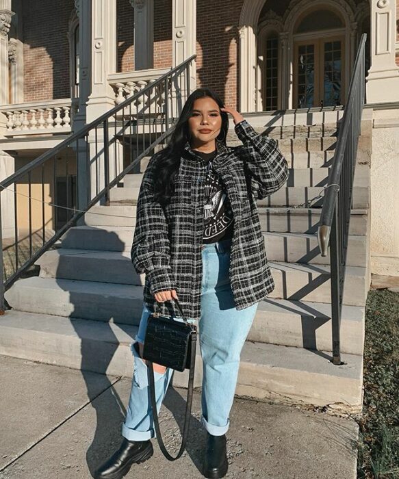 Chica curvy usando botines negros, mom jeans azules, blusa gris y abrigo gris a cuadros, con bolsa de mano negra