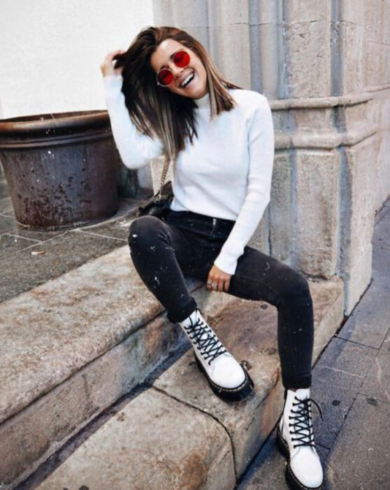 Chica usando botines blancos y jeans negros, con blusa de manga larga blanca y lentes de sol de cristal rojo