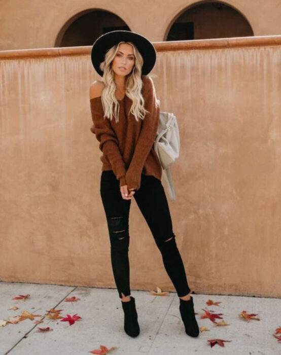 Chica usando sombrero de color negro con outfit de jeans y botines negros, suéter con escote de ojal color camel y bolso de mano blanco