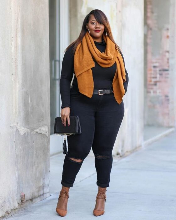 Chica curvy usando jeans y suéter negros, con bufanda mostaza y tacones del mismo color