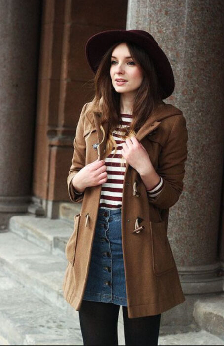 Chica usando sombrero de color negro con outfit de medias negras, mini falda denim, blusa a rayas y abrigo color caki