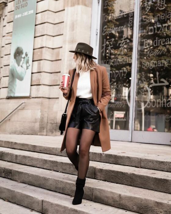 Outfit para la oficina de otoño-invierno de abrigo color camel, suéter blanco, shorts negros, medias y botines negros, además de sombrero negro