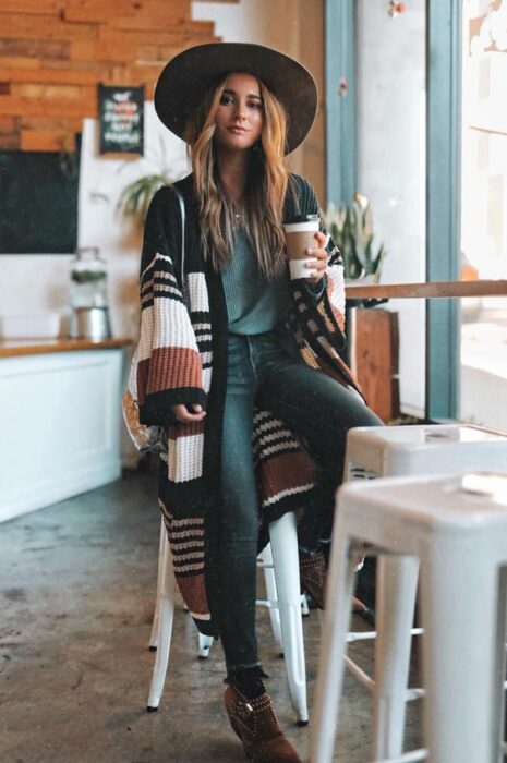 Chica usando sombrero de color negro con outfit de jeans, blusa azul petróleo y cardigan largo de rayas de diferentes colores 