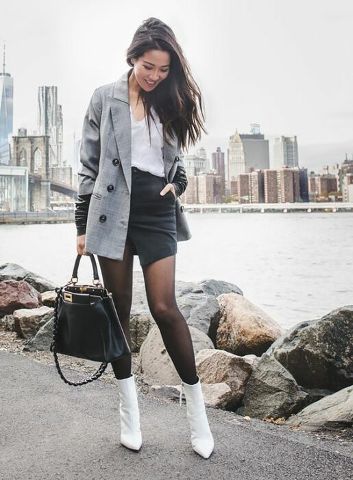 Chica usando botines blancos y medias negras, mini falda de vinil, y blusa blanca además de blazer gris con bolso de mano negro