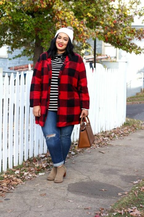 Chica curvy usando jeans rasgados, botines camel, blusa negra con líneas blancas y chaqueta color rojo con cuadros negros