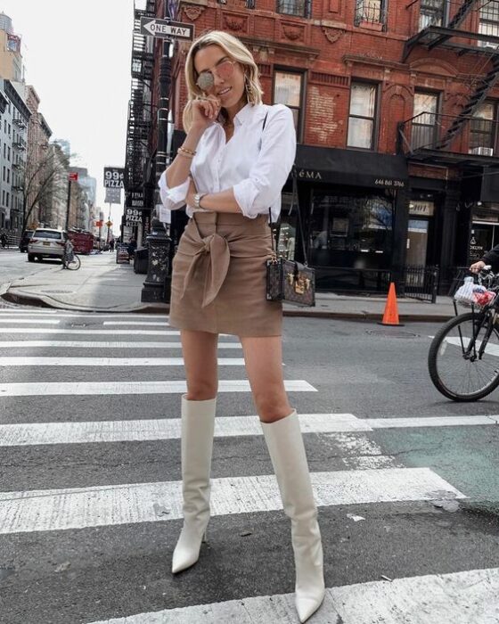 Chica usando botas blancas y mini falda color caki, con camisa color blanca de manga tres cuartos