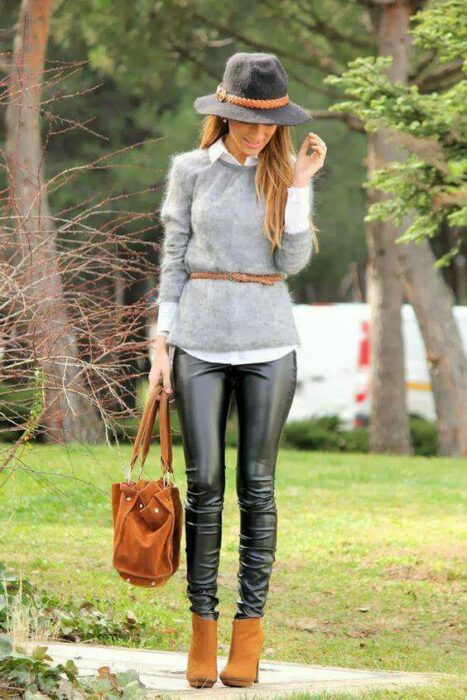 Chica usando sombrero de color gris con outfit de botines color camel, leggings de vinil, camisa blanca con un suéter gris y cinturón camel