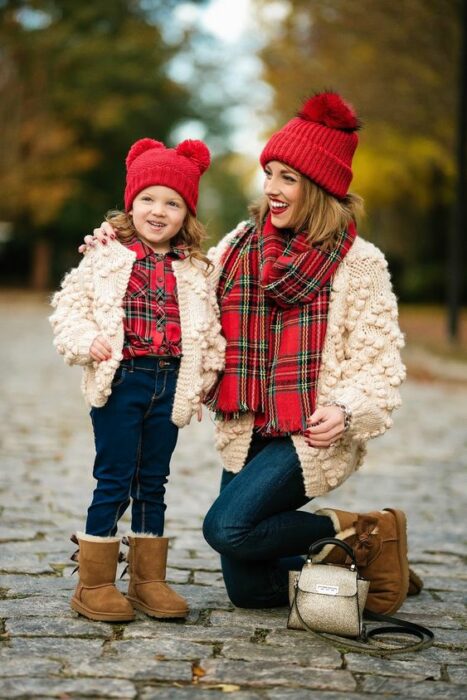 Madre e hija pequeña ambas vistiendo gorrito rojo, camisa tartán roja con verde, suéter blanco con bolitas, jeans oscuros y botas estilo ugg color café