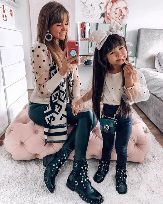 Madre de cabello rubio sentada en un cojín grande rosa y su hija de cabello largo, negro y lacio se toman selfie frente al espejo ambas vistiendo mallones negros, botines negros con aplicaciones blancas y plateadas y una blusa blanca con puntos negros con transparencias en las mangas