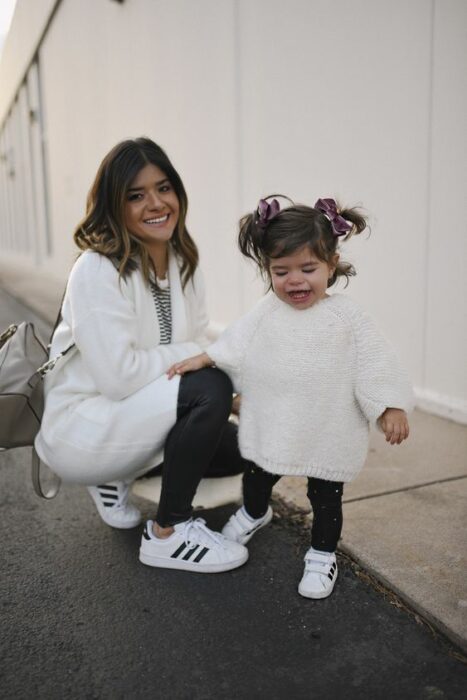 Madre inclinada vistiendo jeans oscuros, tenis blancos con rayas negras, blusa de rayas blancas con negra y un cardigan blanco sostiene a su hija pequeña peinada con dos colitas altas atadas con moños morados de terciopelo mientras viste suéter blanco largo, mallones negros y tenia blancos 