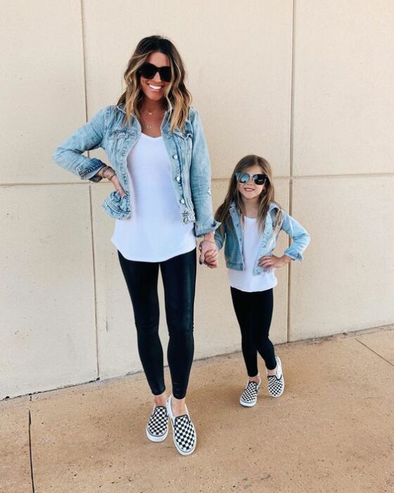 Madre e hija de cabello rubio tomadas de la mano mientras visten mallones negros, vans blancos con cuadros negros, blusa blanca larga, chaqueta de mezclilla y gafas de sol oscuras y tienen la mano en la cintura