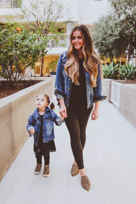 Madre e hija pequeña tomadas de la mano caminando por la calle vistiendo outfits iguales con vestido y pantalón negros, chaqueta de mezclilla oscura y zapatos en 'animal print'