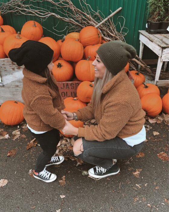 Madre e hija pequeña en pumpkin patch tomadas de las manos con outfits iguales con pantalón negro, converse negros, blusa blanca, abrigo estilo peluche café y gorrito negra y verde