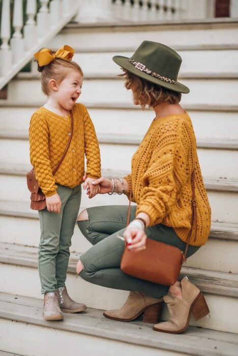 Madre e hija sonriendo en escalones blancos ambas con atuendo iguales de pantalón verde oliva, botines beige, suéter tejido amarillo mostaza, bolso camel mientas que la niña usa un moño grande amarillo y la madre un sombrero verde olivo  