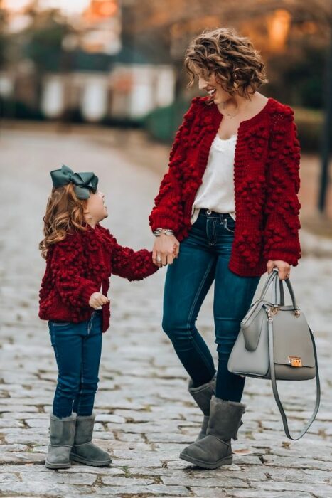 Madre e hija tomadas de la mano caminando en la calle ambas con botas grises UGG, jeans azul marino, blusa blanca y abrigo rojo con bolitas. La niña usa un moño grande verde olivo en el cabello y la madre lleva un bolso gris en la mano izquierda