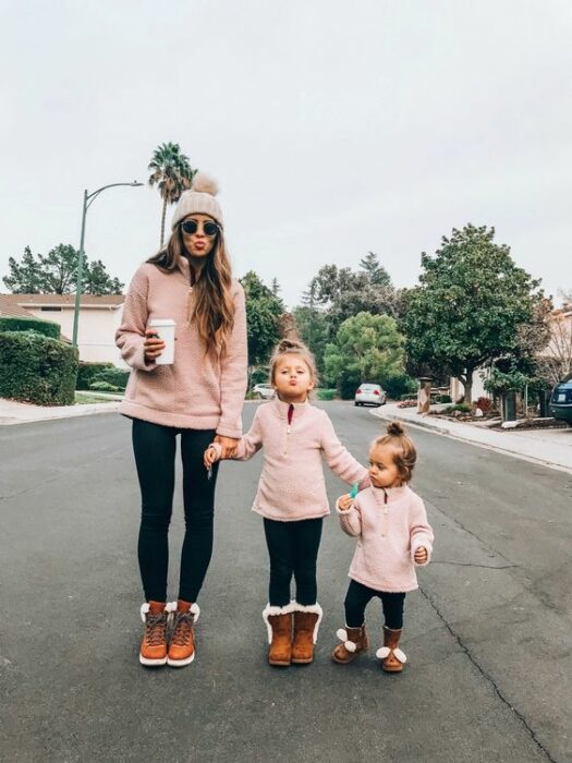 Madre y sus dos hijas visten mallones negros, botas cafés con blanco y sudadera rosa pastel. La madre sostiene un vaso de café blanco, mientras usa gafas de sol negras y un gorro rosa