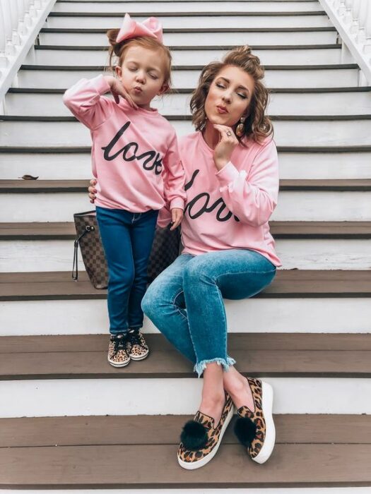 Madre e hija pequeña hacen 'duck face' en escalones blancos mientras ambas visten jeans oscuros, tenis en 'animal print', sudadera rosa que dice la palabra love en letras cursivas y la niña tiene un chongo alto con un moño grande rosa y la madre el cabello corto rizado con un pequeño chongo en la parte superior