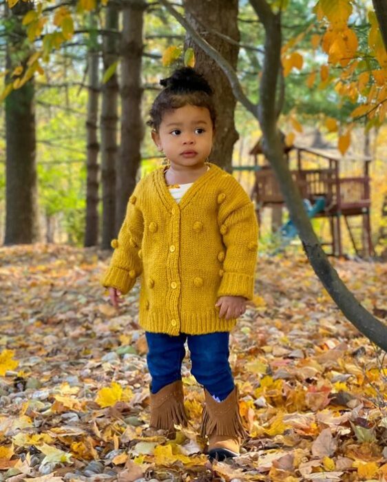 Niña pequeña de cabello rizado peinado en un chongo alto vistiendo suéter amarillo, jeans oscuros y botitas cafés