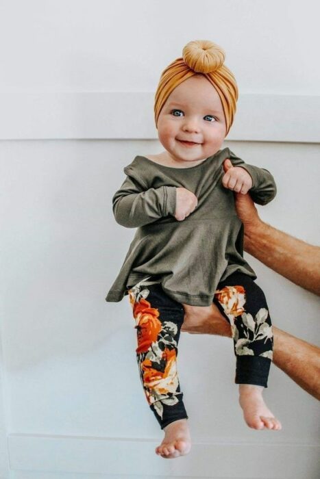 Bebé cargada en brazos con vestidito verde y leggins negros con flores naranjas 