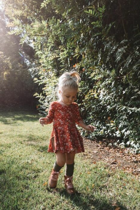 Niña rubia con coleta alta vistiendo vestido floreado color naranja con calcetas negras y botas cafés 