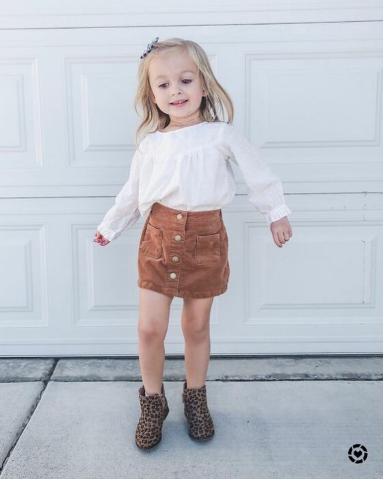 Niña pequeña rubia con blusa blanca, falda naranja de pana y botas cafés 