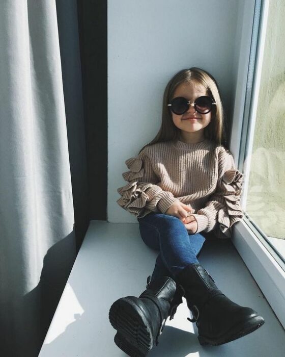 Niña De cabello castaño largo sentada en la ventana vistiendo suéter beige, pantalón de mezclilla, botas engras altas y lentes negros de sol