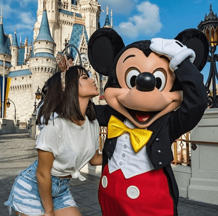 Chica de melena corta café con blusa blanca y shorts de mezclilla besando a Mickey MOuse