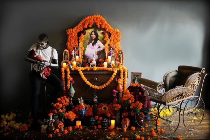 Hombre vestido de catrín posa en altar de muertos que hizo en honor a su esposa mientras sostiene a su bebé en brazos y la mira