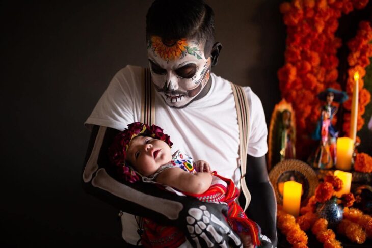 Hombre vestido de catrín posa en altar de muertos que hizo en honor a su esposa mientras sostiene a su bebé en brazos