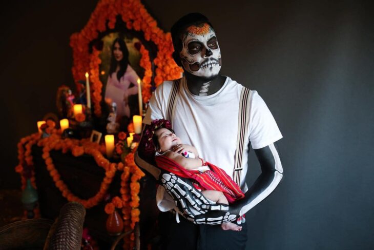 Hombre vestido de catrín posa en altar de muertos que hizo en honor a su esposa mientras sostiene a su bebé en brazos y mira por la ventana