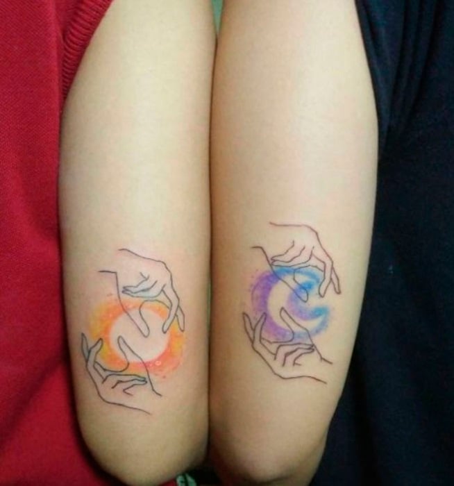 Tatuaje complementario de un sol y una luna con dos manos al rededor de ellos, sobre el antebrazo