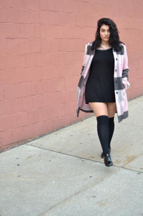 Chica curvy usando un vestido color negro con botas altas y camisa de cuadros color gris y rosa