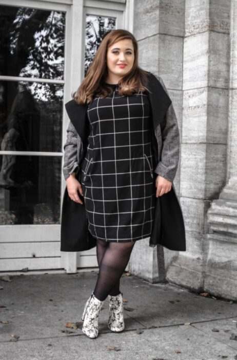 Chica curvy usando un vestido color negro con cuadros blancos, blazer gris, medias negras y botas pitón 