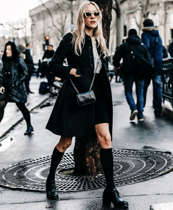 chica rubia usando lentes de sol, camisa de vestir blanca, abrigo negro largo, minifalda negra, calcetas negras largas, zapatos de plataforma negros y bolso mini negro